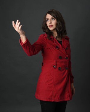 Güzel esmer kadın modelinin yakın plan portresi, kırmızı trençkot giyiyor, el hareketleriyle poz veriyor. Karanlık stüdyo arka planında gölgeler ile izole edilmiş.. 