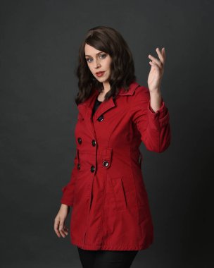 Güzel esmer kadın modelinin yakın plan portresi, kırmızı trençkot giyiyor, el hareketleriyle poz veriyor. Karanlık stüdyo arka planında gölgeler ile izole edilmiş.. 