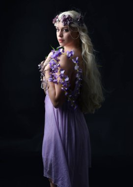 Uzun sarı saçlı, mor peri elbisesi giyen ve fantezi çiçekli taç takan güzel bir kadın modelin portresi. Stüdyonun arka planında izole edilmiş, elinde mor çiçekler var.