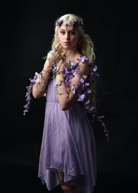 Uzun sarı saçlı, mor peri elbisesi giyen ve fantezi çiçekli taç takan güzel bir kadın modelin portresi. Stüdyonun arka planında izole edilmiş, elinde mor çiçekler var.