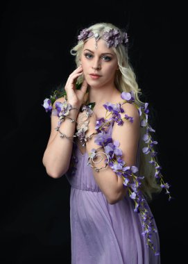 Uzun sarı saçlı, mor peri elbisesi giyen ve fantezi çiçekli taç takan güzel bir kadın modelin portresi. Stüdyonun arka planında izole edilmiş, elinde mor çiçekler var.
