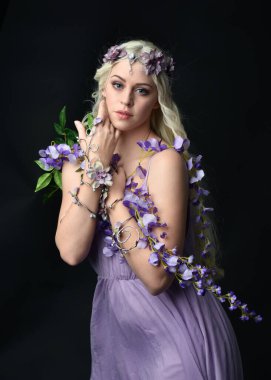 Uzun sarı saçlı, mor peri elbisesi giyen ve fantezi çiçekli taç takan güzel bir kadın modelin portresi. Stüdyonun arka planında izole edilmiş, elinde mor çiçekler var.