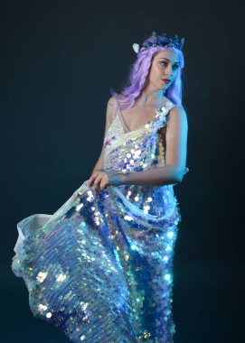 Uzun mor saçlı, fantezi tacını takan, gökkuşağı pullu balo elbisesi giyen güzel bir kadın modelin sanatsal portresi. Karanlık stüdyo arka planında izole edilmiş hareketli duruş..