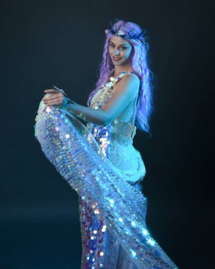 Uzun mor saçlı, fantezi tacını takan, gökkuşağı pullu balo elbisesi giyen güzel bir kadın modelin sanatsal portresi. Karanlık stüdyo arka planında izole edilmiş hareketli duruş..