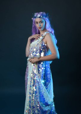 Uzun mor saçlı, fantezi tacını takan, gökkuşağı pullu balo elbisesi giyen güzel bir kadın modelin sanatsal portresi. Karanlık stüdyo arka planında izole edilmiş hareketli duruş..