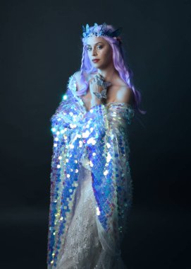 Uzun mor saçlı, fantezi tacını takan, gökkuşağı pullu balo elbisesi giyen güzel bir kadın modelin sanatsal portresi. Karanlık stüdyo arka planında izole edilmiş hareketli duruş..