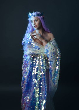 Uzun mor saçlı, fantezi tacını takan, gökkuşağı pullu balo elbisesi giyen güzel bir kadın modelin sanatsal portresi. Karanlık stüdyo arka planında izole edilmiş hareketli duruş..