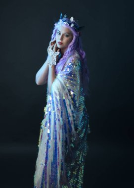 Uzun mor saçlı, fantezi tacını takan, gökkuşağı pullu balo elbisesi giyen güzel bir kadın modelin sanatsal portresi. Karanlık stüdyo arka planında izole edilmiş hareketli duruş..