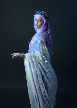 Uzun mor saçlı, fantezi tacını takan, gökkuşağı pullu balo elbisesi giyen güzel bir kadın modelin sanatsal portresi. Karanlık stüdyo arka planında izole edilmiş hareketli duruş..