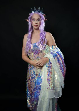 Güzel bir kadın modelin sanatsal portresi, uzun mor saçlı ve elf kulaklı, fantezi bir peri tacı takan, gökkuşağı pullu bir balo elbisesi giyen. Yan profilde duruyor, karanlık stüdyo geçmişinde izole edilmiş..