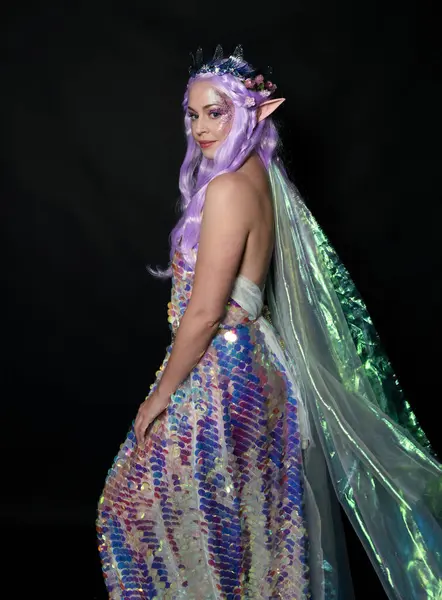 artistic portrait of beautiful female model with long purple hair and elf ears, wearing a fantasy fairy crown, wearing a rainbow glitter sequin ball gown. Standing in side profile, isolated on dark studio background.