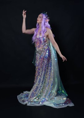 Uzun mor saçlı, elf kulakları takan güzel kadın modelin tam boy portresi, fantezi bir peri tacı ve gökkuşağı pullu balo elbisesi. Karanlık stüdyo arka planında izole edilmiş zarif duruş.