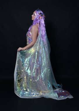 Uzun mor saçlı, elf kulakları takan güzel kadın modelin tam boy portresi, fantezi bir peri tacı ve gökkuşağı pullu balo elbisesi. Uzaklaşmak, arka plan, izole edilmiş karanlık stüdyo 