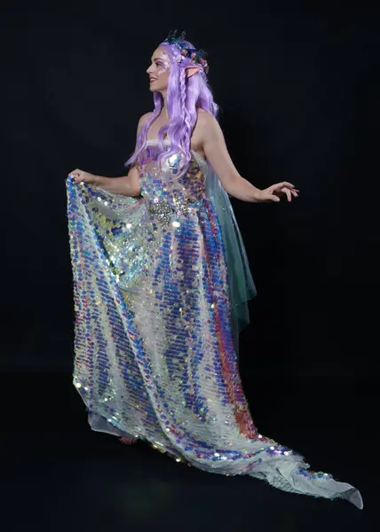 Full length portrait of beautiful female model with long purple hair wearing elf ears, a fantasy fairy crown and  rainbow glitter sequin ball gown. graceful standing pose,  isolated in dark studio background