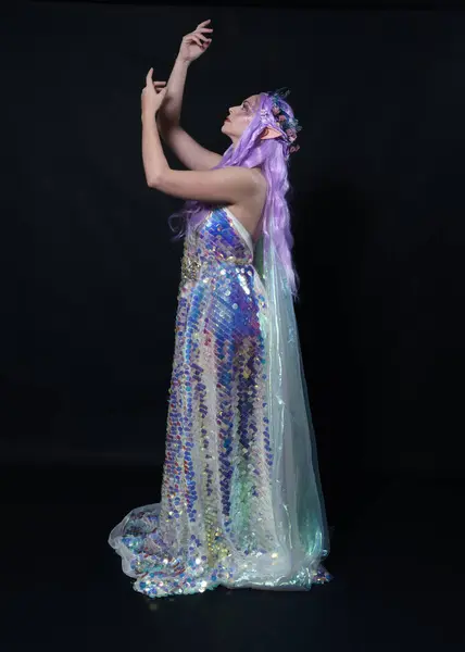 Full length portrait of beautiful female model with long purple hair wearing elf ears, a fantasy fairy crown and  rainbow glitter sequin ball gown.  walking away, back view, isolated dark studio