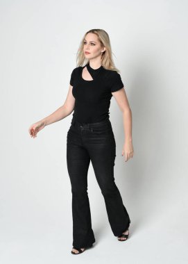 Modern siyah gömlek ve deri pantolon giyen güzel sarışın kadının tam boy portresi. Beyaz stüdyo arka planında izole edilmiş, el hareketleriyle kendine güvenen duruş..