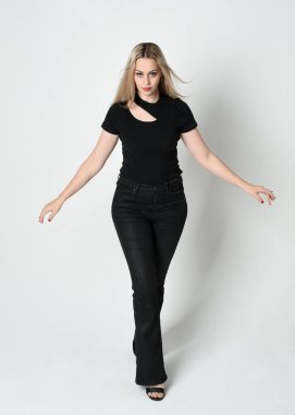 Modern siyah gömlek ve deri pantolon giyen güzel sarışın kadının tam boy portresi. Beyaz stüdyo arka planında izole edilmiş, el hareketleriyle kendine güvenen duruş..