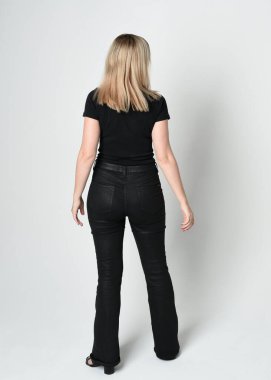 Modern siyah gömlek ve deri pantolon giyen güzel sarışın kadının tam boy portresi. Kameradan uzaklaşan kendine güvenen duruş, beyaz stüdyo arka planında siluet..