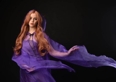 Uzun kızıl saçlı, mor peri fantezi cüppeli güzel bir kadın modelin portresi. Zarif jestsel el büyü yapıyor. Koyu siyah stüdyo arka planında izole.