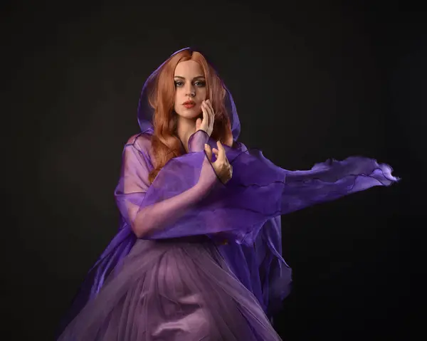 stock image close up portrait of beautiful female model with long red hair, wearing  purple fairy fantasy cloak gown. Elegant gestural hand poses casting magic spell. Isolated on dark black studio background