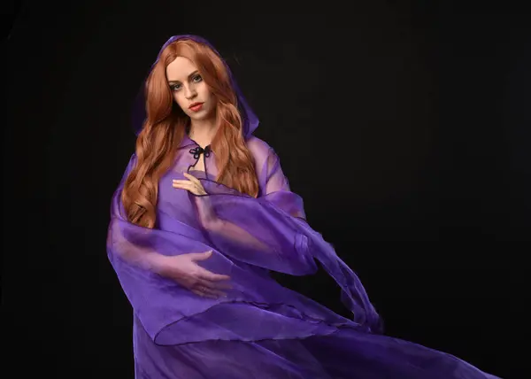 Stock image close up portrait of beautiful female model with long red hair, wearing  purple fairy fantasy cloak gown. Elegant gestural hand poses casting magic spell. Isolated on dark black studio background