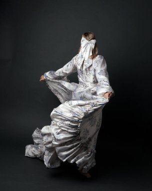 Romantik tarihi gelinlik giyen güzel sarışın kadın modelin portresi beyaz çiçek desenli. Ayakta duran poz kameradan süzülen ipek etekle kaçıyor. izole edilmiş karanlık stüdyo.