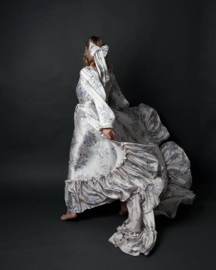 Romantik tarihi gelinlik giyen güzel sarışın kadın modelin portresi beyaz çiçek desenli. Ayakta duran poz kameradan süzülen ipek etekle kaçıyor. izole edilmiş karanlık stüdyo.