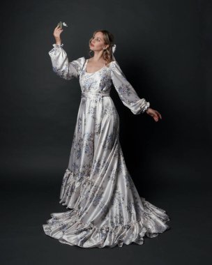 Romantik tarihi gelinlik giyen güzel sarışın kadın modelin portresi beyaz çiçek desenli. Dalgalı ipek etekli duruş. izole edilmiş karanlık stüdyo.