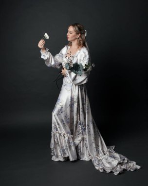 Romantik tarihi gelinlik giyen güzel sarışın kadın modelin portresi beyaz çiçek desenli. Dalgalı ipek etekli duruş. izole edilmiş karanlık stüdyo.