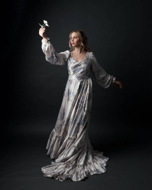 Romantik tarihi gelinlik giyen güzel sarışın kadın modelin portresi beyaz çiçek desenli. Dalgalı ipek etekli duruş. izole edilmiş karanlık stüdyo.