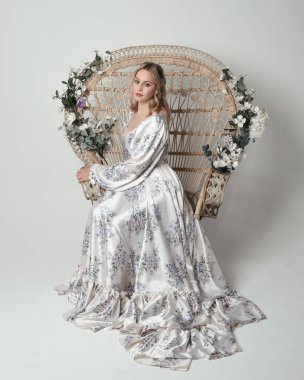 Romantik tarihsel beyaz gelinlik giyen güzel sarışın kadın modelin portresi. Çiçekli tavus kuşu tahtına oturmuş ipek etekle poz veriyor. izole edilmiş karanlık stüdyo.