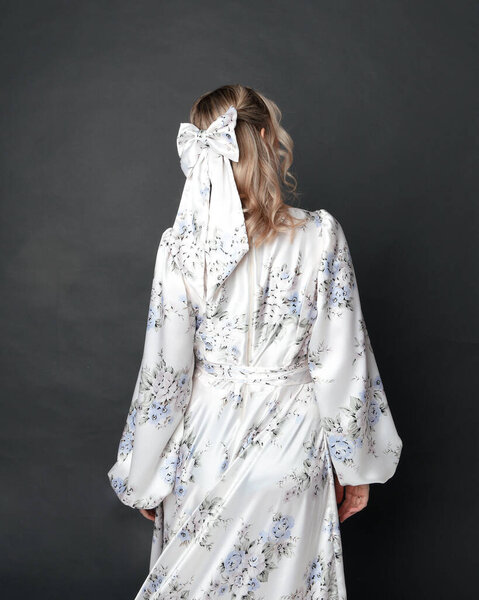 close up portrait of beautiful blonde female model wearing romantic historical gown of white silk with floral pattern and bow. Elegant standing pose, isolated on dark studio background.