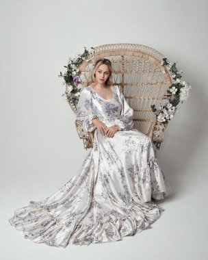 Romantik tarihsel beyaz gelinlik giyen güzel sarışın kadın modelin portresi. Çiçekli tavus kuşu tahtına oturmuş ipek etekle poz veriyor. izole edilmiş karanlık stüdyo.