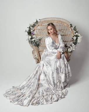 Romantik tarihsel beyaz gelinlik giyen güzel sarışın kadın modelin portresi. Çiçekli tavus kuşu tahtına oturmuş ipek etekle poz veriyor. izole edilmiş karanlık stüdyo.