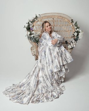 Romantik tarihsel beyaz gelinlik giyen güzel sarışın kadın modelin portresi. Çiçekli tavus kuşu tahtına oturmuş ipek etekle poz veriyor. izole edilmiş karanlık stüdyo.