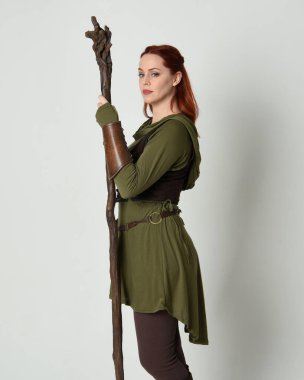  Yeşil ortaçağ fantezi kostümü ve kahverengi zırhlı güzel kızıl saçlı kadın modelin portresi. Tahta sihirbaz silahıyla ayakta duruyor, izole edilmiş beyaz stüdyo arka planı..