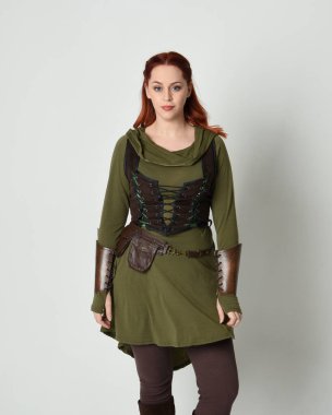 Yeşil ve kahverengi ortaçağ fantezi kostümlü, tunik ve zırhlı güzel kızıl saçlı kadın modelin yakın portresi. Beyaz stüdyo arka planında izole edilmiş, el hareketleriyle duruyor..