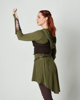 Yeşil ve kahverengi ortaçağ fantezi kostümlü, tunik ve zırhlı güzel kızıl saçlı kadın modelin yakın portresi. Beyaz stüdyo arka planında izole edilmiş, el hareketleriyle duruyor..