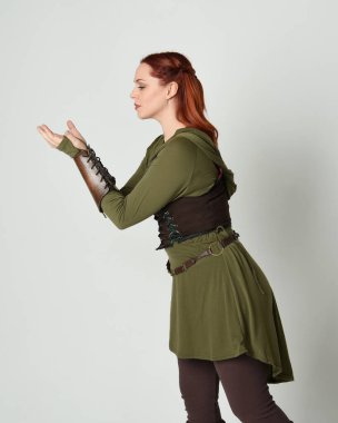 Yeşil ve kahverengi ortaçağ fantezi kostümlü, tunik ve zırhlı güzel kızıl saçlı kadın modelin yakın portresi. Beyaz stüdyo arka planında izole edilmiş, el hareketleriyle duruyor..