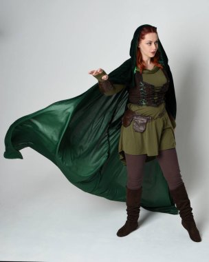 Yeşil ortaçağ fantezi deri zırh giyen kızıl saçlı kadın modelin tam boy portresi, dramatik uzun kadife kapüşonlu pelerin. Yürüyen poz, izole edilmiş beyaz stüdyo arka planı..