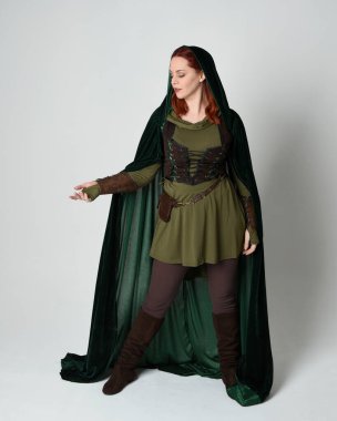 Yeşil ortaçağ fantezi deri zırh giyen kızıl saçlı kadın modelin tam boy portresi, dramatik uzun kadife kapüşonlu pelerin. Yürüyen poz, izole edilmiş beyaz stüdyo arka planı..