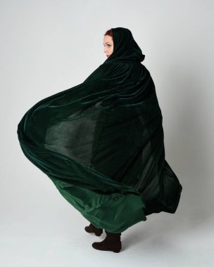 Kızıl saçlı kadın modelin tam boy portresi, yeşil ortaçağ fantezi kostümü, uzun kadife kapüşonlu pelerin. Duruş, geri geri yürüme, izole beyaz stüdyo arka planı.