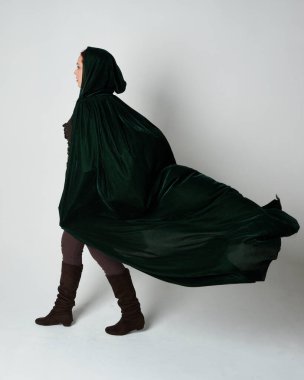 Kızıl saçlı kadın modelin tam boy portresi, yeşil ortaçağ fantezi kostümü, uzun kadife kapüşonlu pelerin. Duruş, geri geri yürüme, izole beyaz stüdyo arka planı.