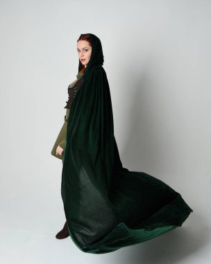 Kızıl saçlı kadın modelin tam boy portresi, yeşil ortaçağ fantezi kostümü, uzun kadife kapüşonlu pelerin. Duruş, geri geri yürüme, izole beyaz stüdyo arka planı.