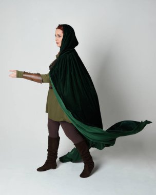 Kızıl saçlı kadın modelin tam boy portresi, yeşil ortaçağ fantezi kostümü, uzun kadife kapüşonlu pelerin. Duruş, geri geri yürüme, izole beyaz stüdyo arka planı.