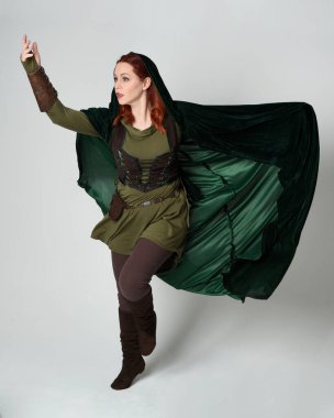 Yeşil ortaçağ fantezi deri zırh giyen kızıl saçlı kadın modelin tam boy portresi, dramatik uzun kadife kapüşonlu pelerin. Yürüyen poz, izole edilmiş beyaz stüdyo arka planı..