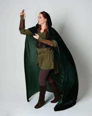 Yeşil ortaçağ fantezi deri zırh giyen kızıl saçlı kadın modelin tam boy portresi, dramatik uzun kadife kapüşonlu pelerin. Yürüyen poz, izole edilmiş beyaz stüdyo arka planı..