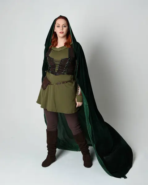 stock image Full length portrait of red haired female model wearing green medieval fantasy costume leather armour, dramatic long flowing hooded velvet cape. Standing walking pose, isolated white studio background.