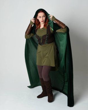 Yeşil ortaçağ fantezi deri zırh giyen kızıl saçlı kadın modelin tam boy portresi, dramatik uzun kadife kapüşonlu pelerin. Yürüyen poz, izole edilmiş beyaz stüdyo arka planı..