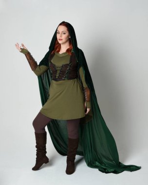 Yeşil ortaçağ fantezi deri zırh giyen kızıl saçlı kadın modelin tam boy portresi, dramatik uzun kadife kapüşonlu pelerin. Yürüyen poz, izole edilmiş beyaz stüdyo arka planı..